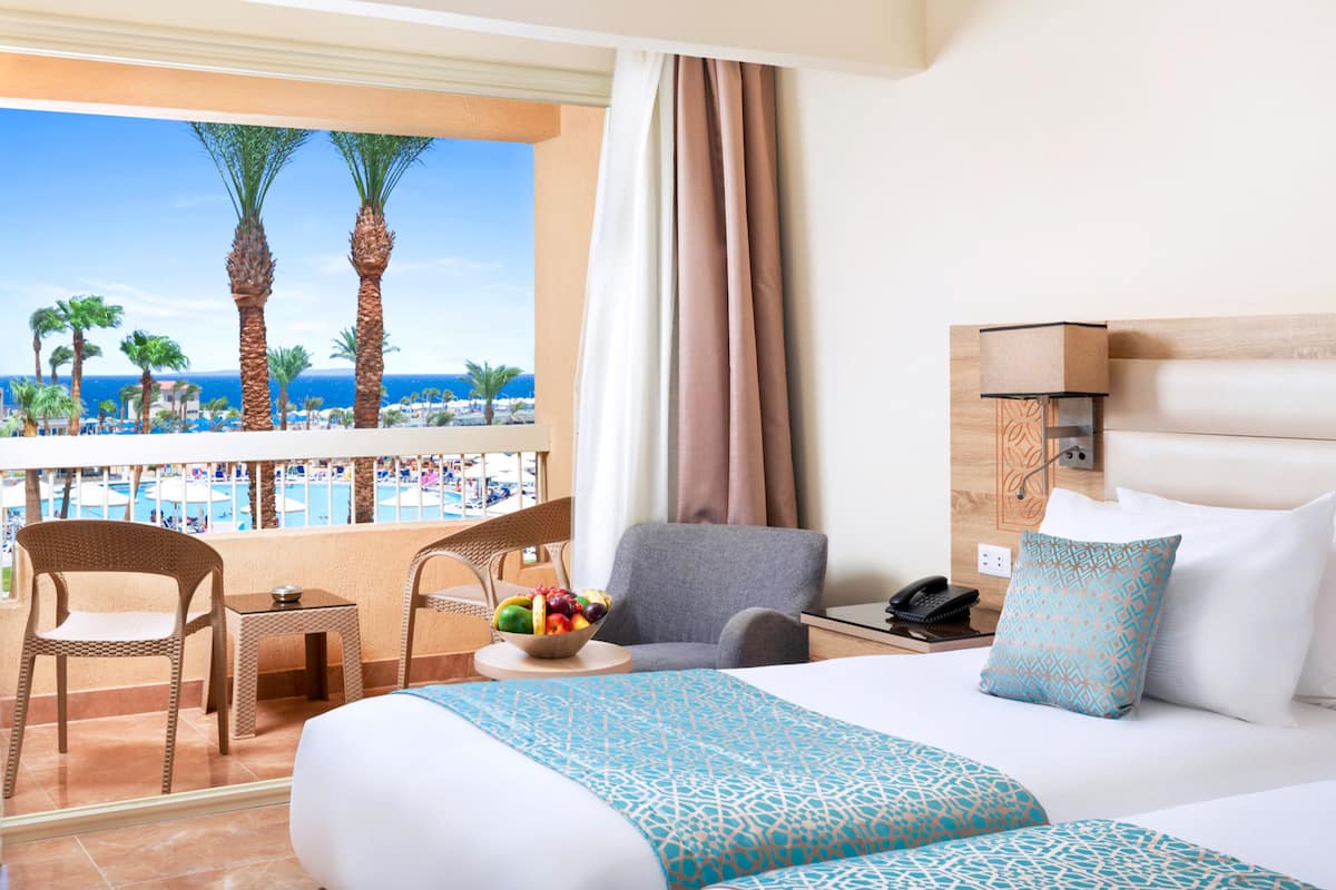 Une chambre d’hôtel avec un balcon, un lit avec une literie à motifs, une chaise, une petite table avec un panier de fruits et un téléphone.