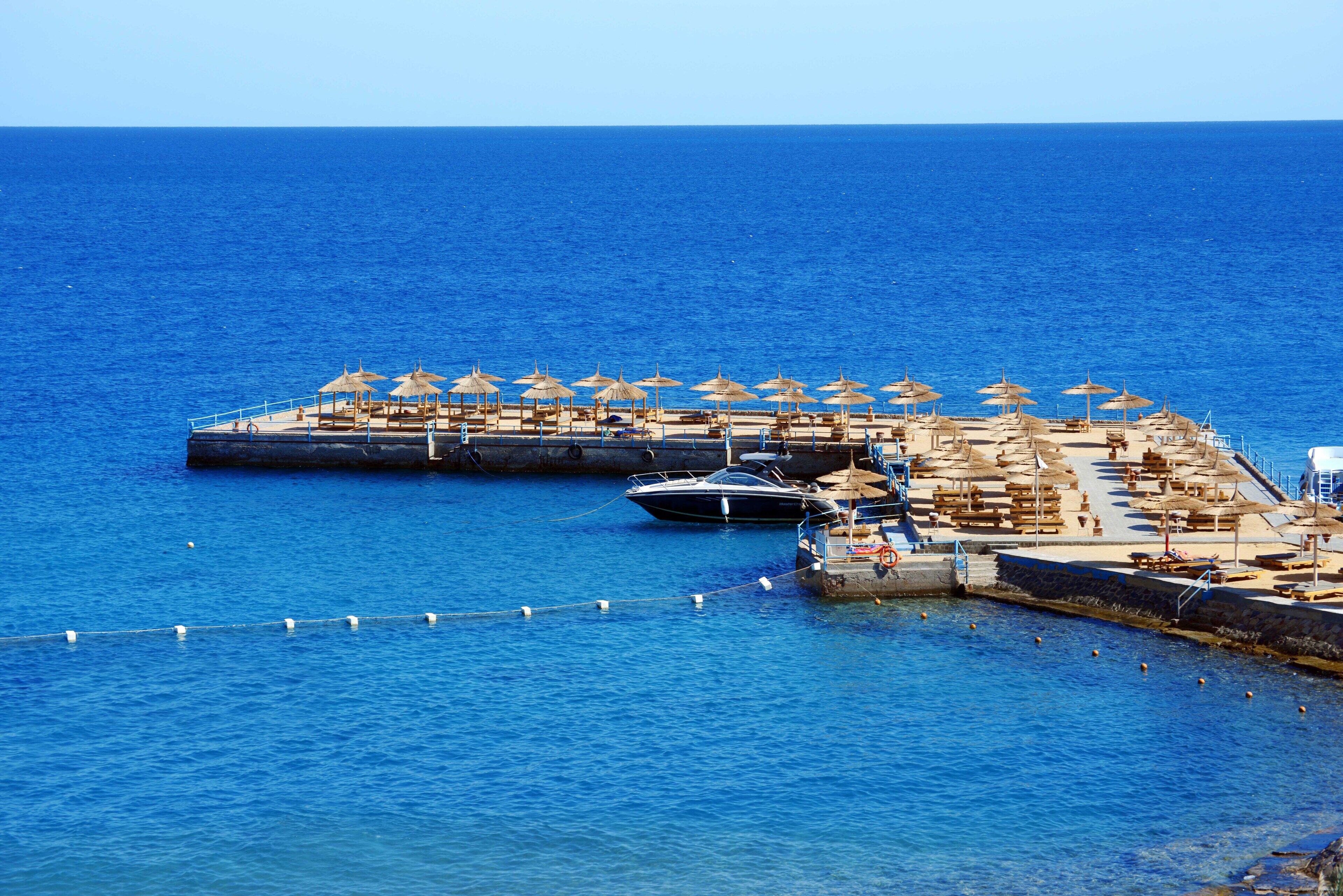 A pier with umbrellas and lounge chairs, boats docked, and a clear blue sea.