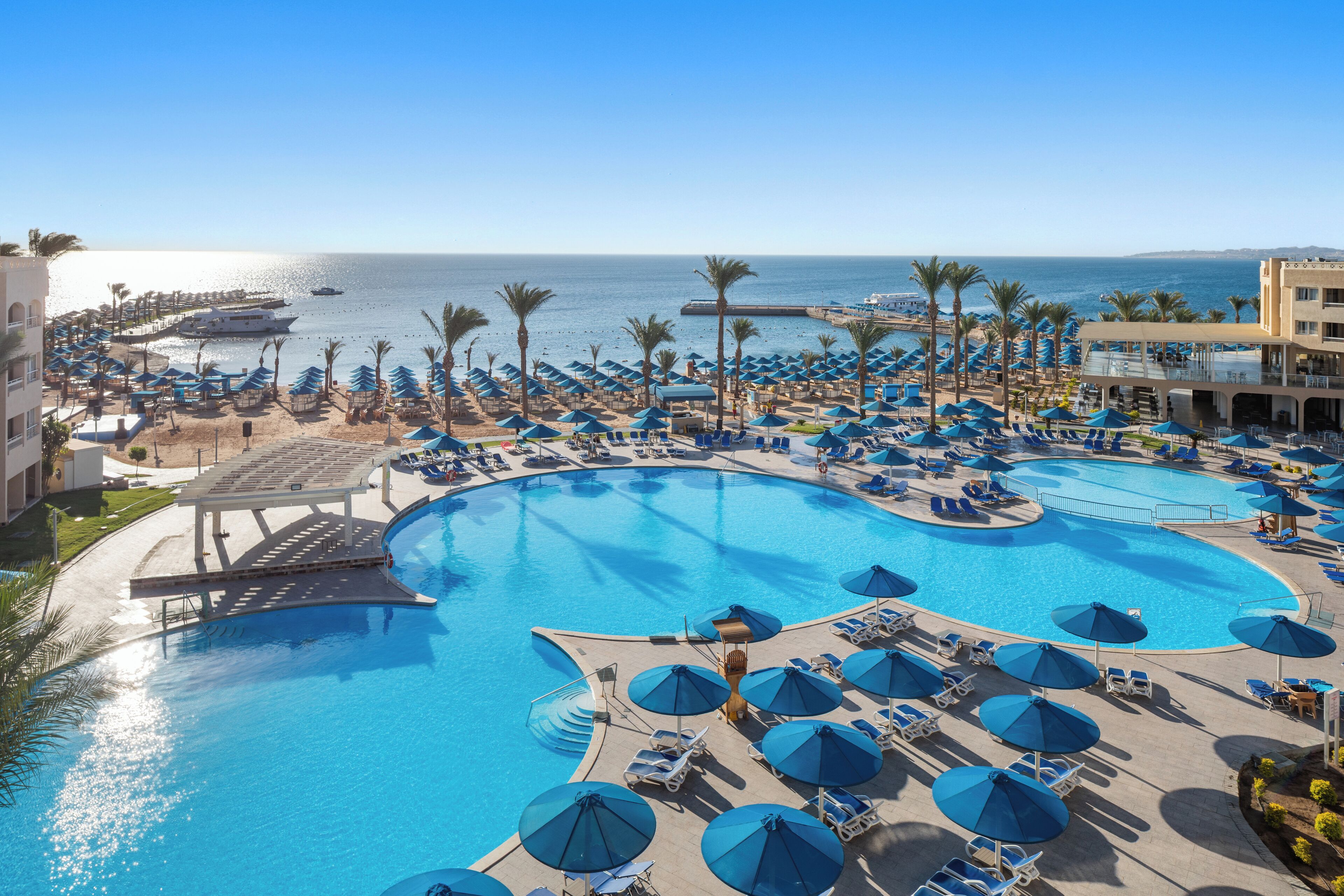 Área de piscina do resort com várias espreguiçadeiras e guarda-sóis, vista clara do oceano e uma piscina grande.