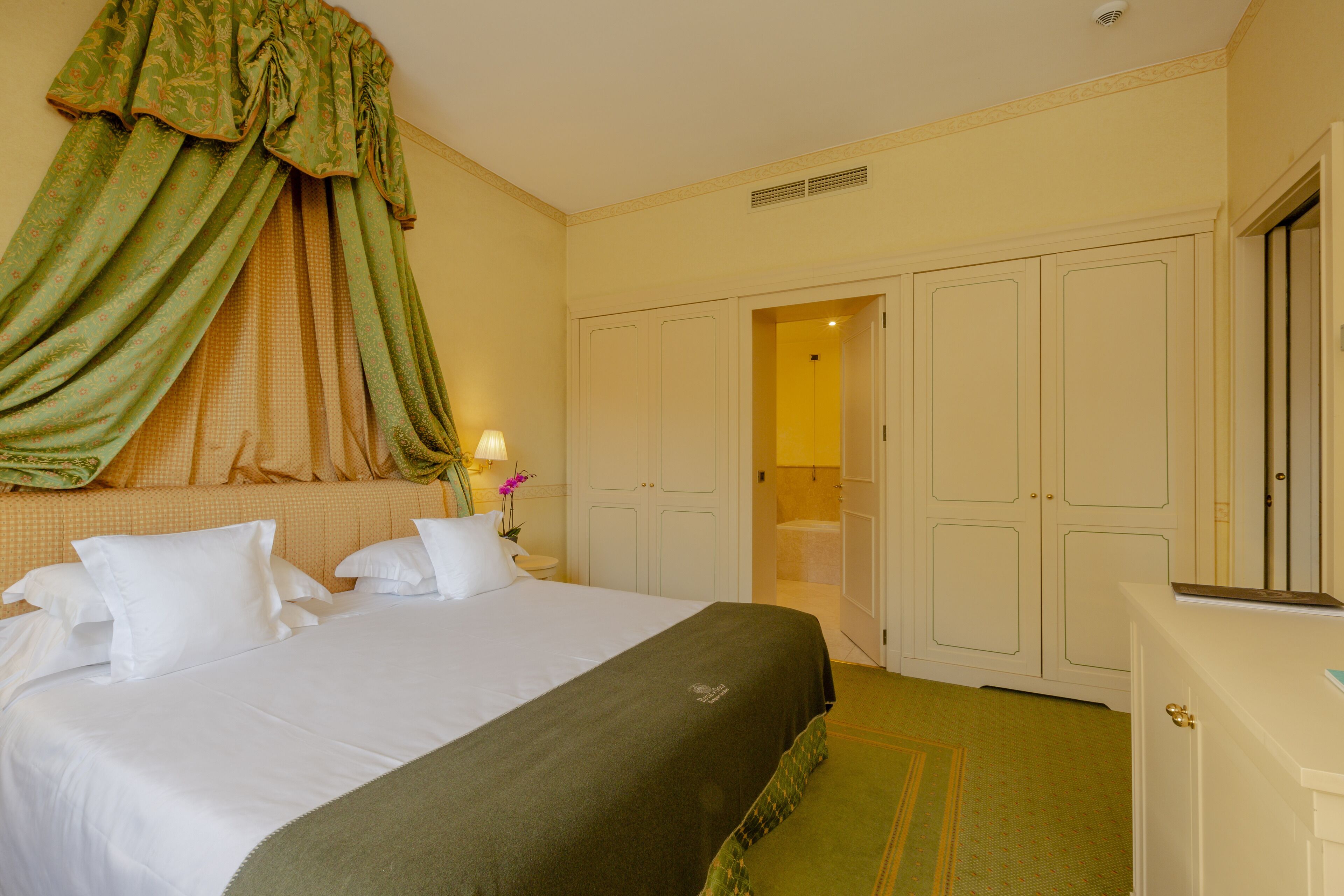A hotel room with a large bed, a green curtain, a white wardrobe, and a small table with a lamp.