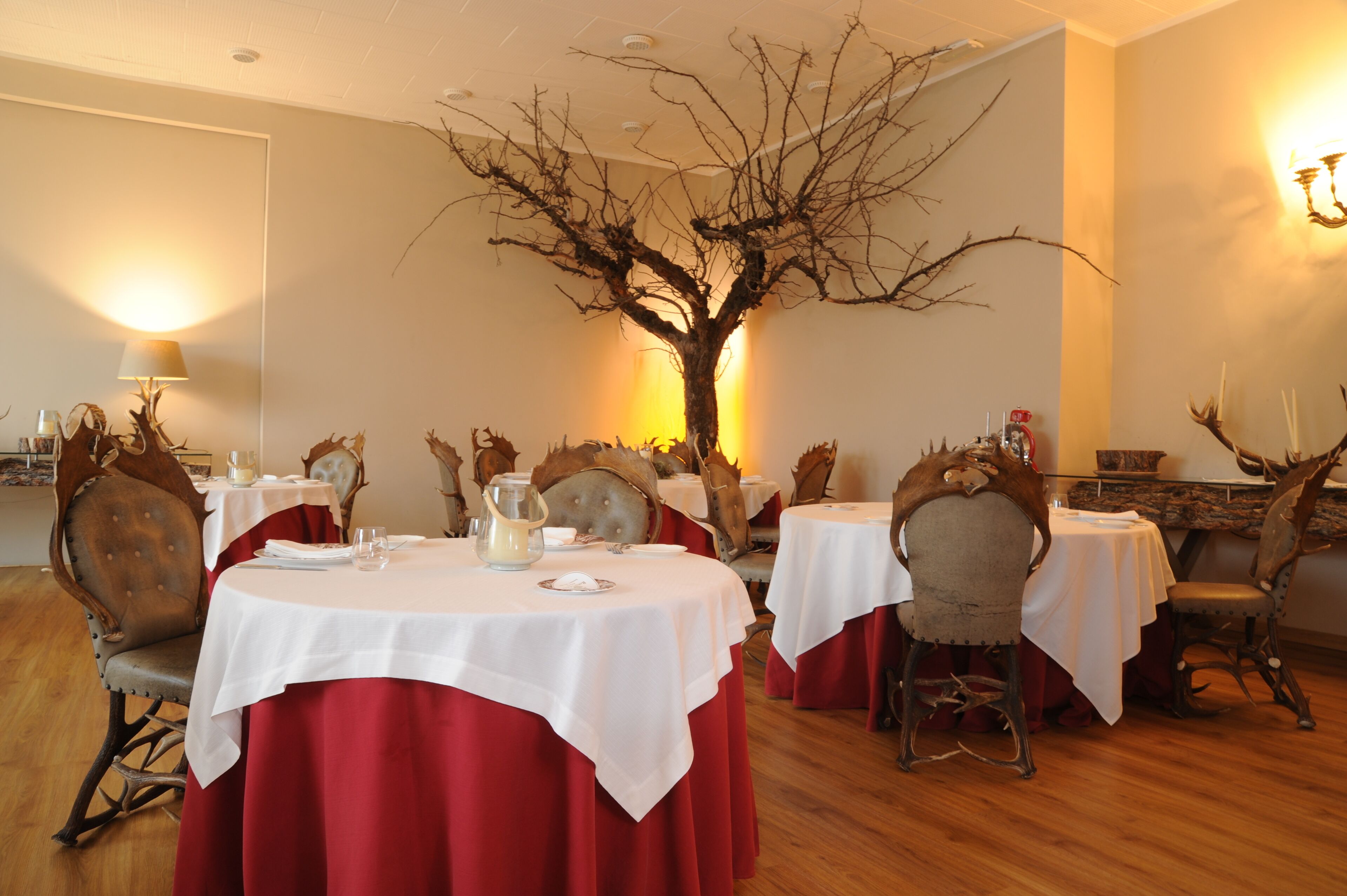 A dining area with round tables covered in white tablecloths and red table runners, surrounded by wooden chairs with antler decorations.