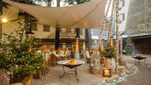 An outdoor seating area with a fire pit, wooden tables, and chairs, surrounded by stone walls and trees.