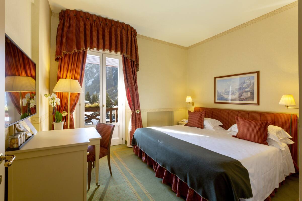 A hotel room with a bed, two bedside lamps, a desk with a lamp, a chair, and a window with curtains.