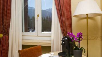 A hotel room with a round table, two chairs, a lamp, a coffee maker, a potted plant, and a window with curtains.