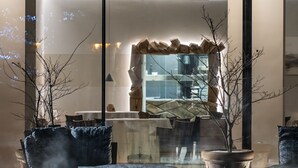 A modern lounge area with a sofa, armchairs, and a coffee table. There are potted plants and a large mirror reflecting the room.