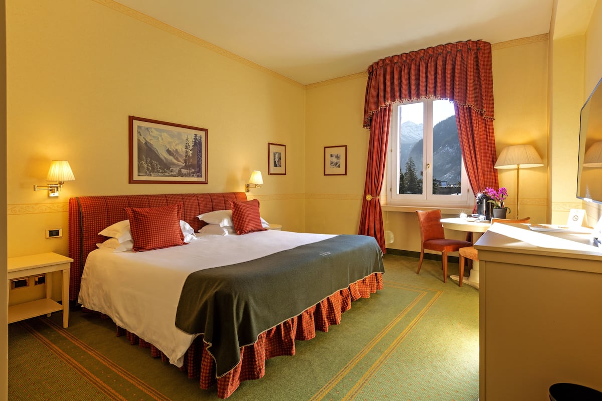 A hotel room with a large bed, a desk, and a chair. There are two lamps on the nightstands and a window with curtains.