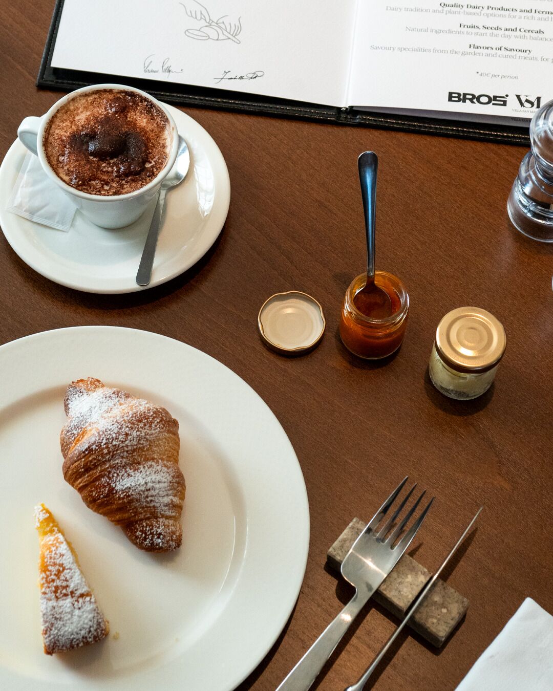A table with a croissant, a chocolate dessert, a spoon, a jar of jam, and a menu.