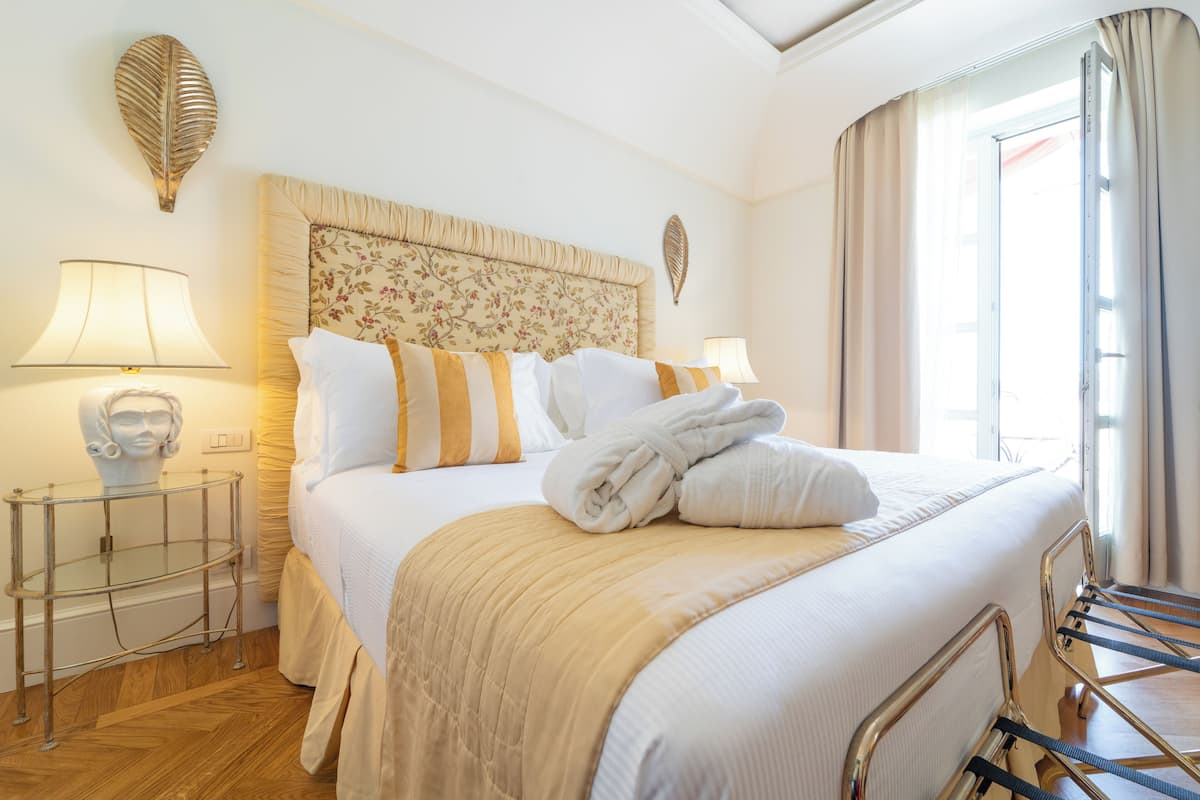 A neatly made bed with a headboard, two bedside lamps, a glass side table, and a window with curtains.