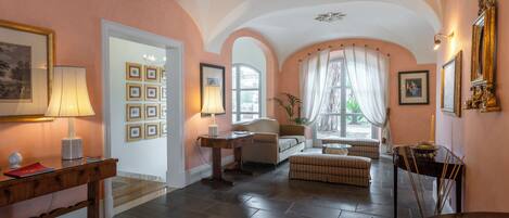 Een ruime kamer met een hoge plafond, een bank, een tafel en een raam met gordijnen.