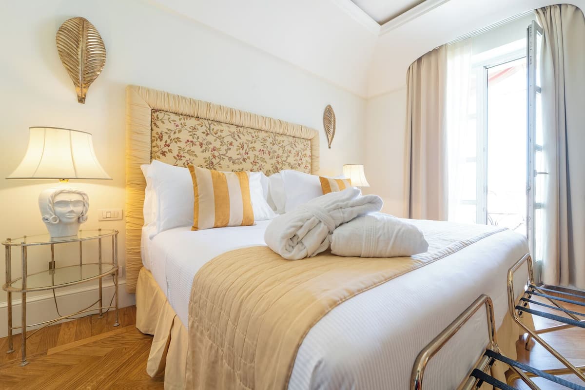 A hotel room with a bed, bedside lamps, a decorative headboard, a glass side table, and a view of the outdoors.
