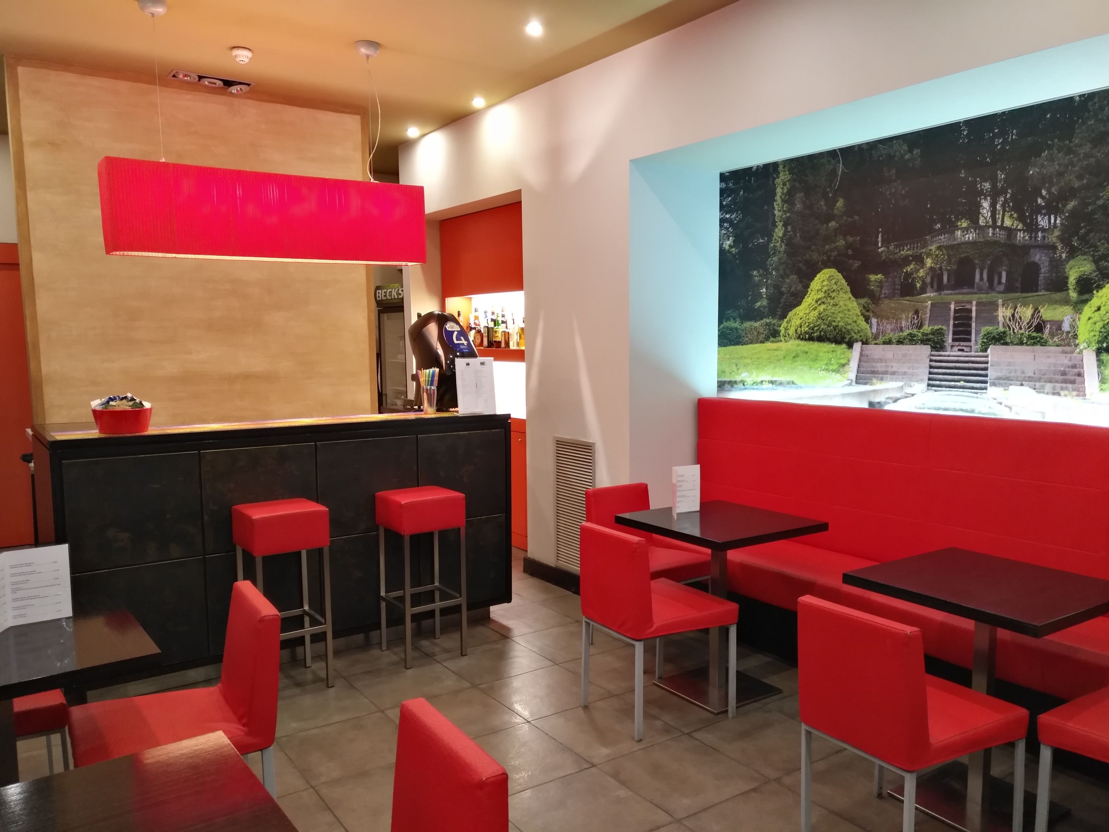 Un restaurante con asientos rojos, una zona de bar y un ventanal con vistas a un jardín.