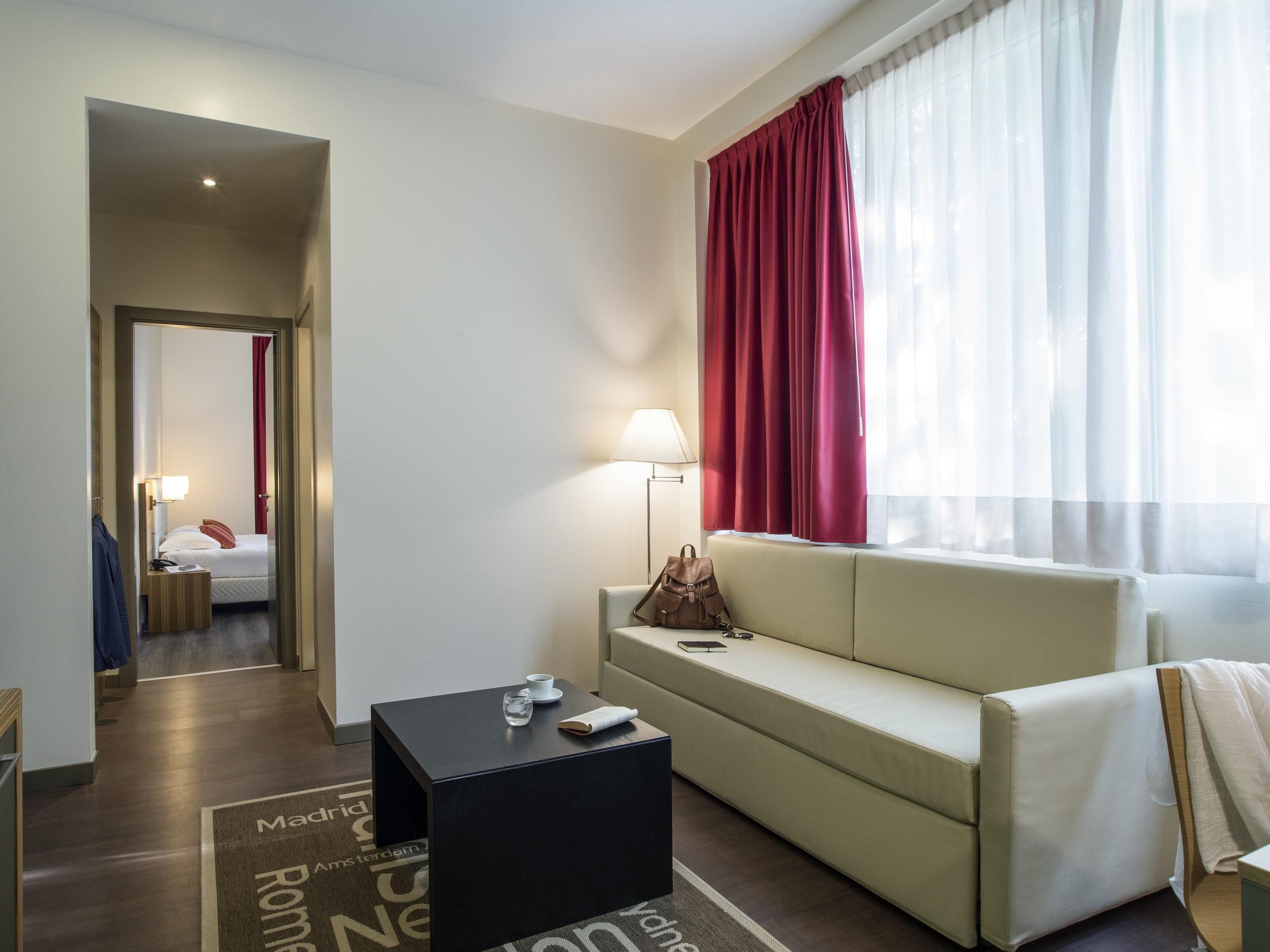 Habitación de hotel con cama, sofá, mesa de centro, lámpara y ventana con cortinas.