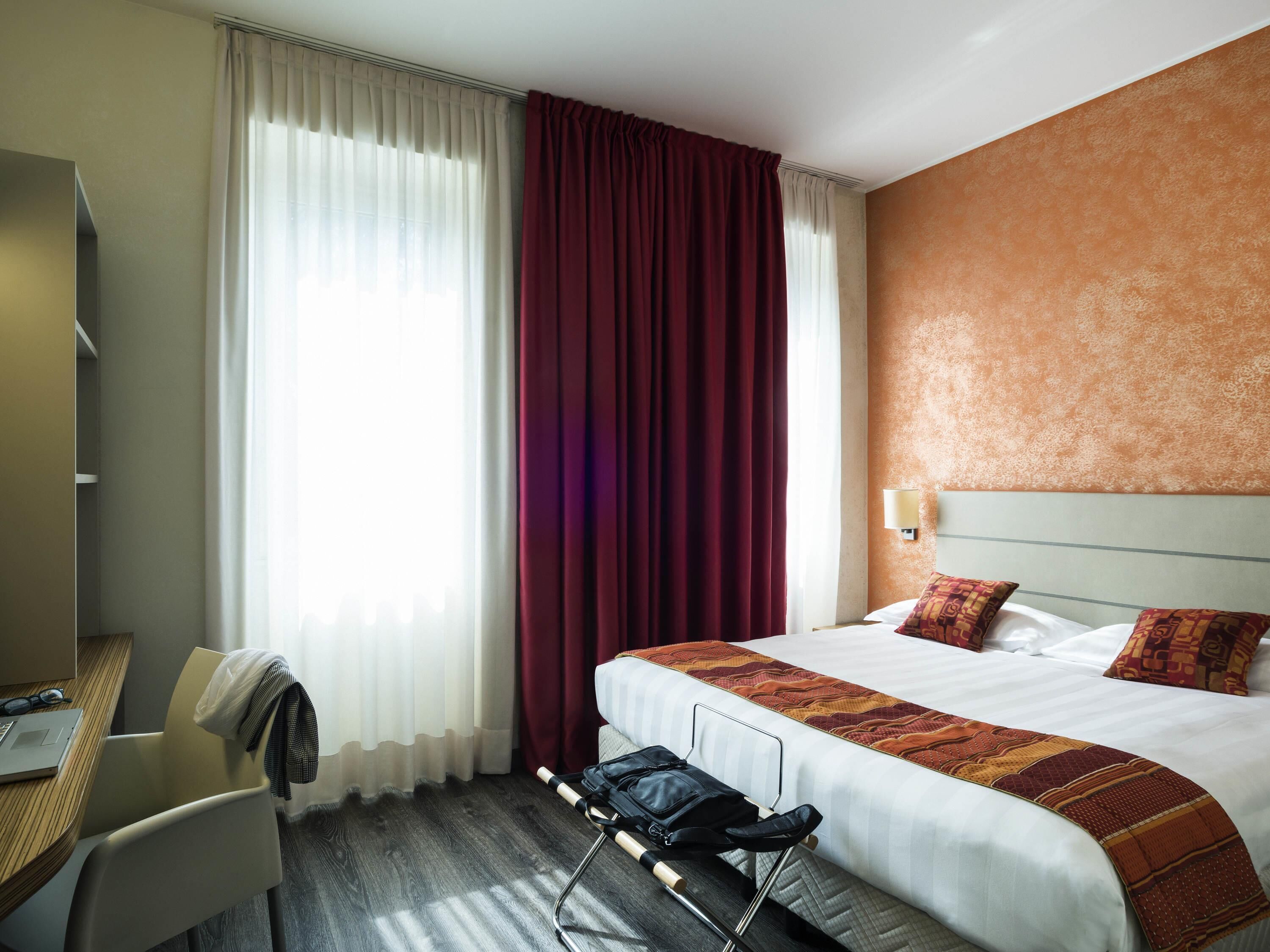Une chambre d’hôtel avec un lit, un bureau, une chaise, une valise et une fenêtre avec des rideaux.