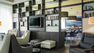 A modern hotel room with a living area, a TV, a bookshelf, and a dining area with a view.