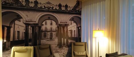 Habitación de hotel con un mural de un patio interior, un sofá, una silla, una mesita y una lámpara.