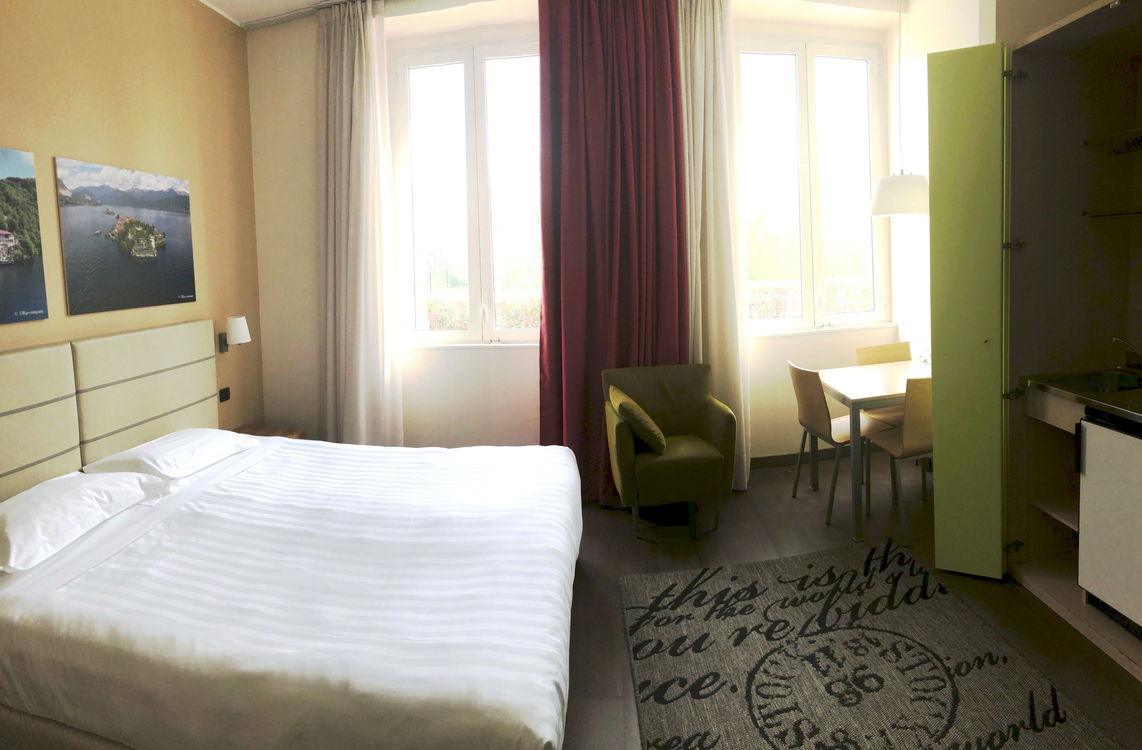 Ein Hotelzimmer mit Bett, Schreibtisch, Stuhl, einem Fenster mit Vorhängen und einem Bild an der Wand.