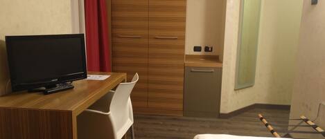 A hotel room with a wooden desk, a television, a white chair, a bed, a wardrobe, and a small kitchenette.