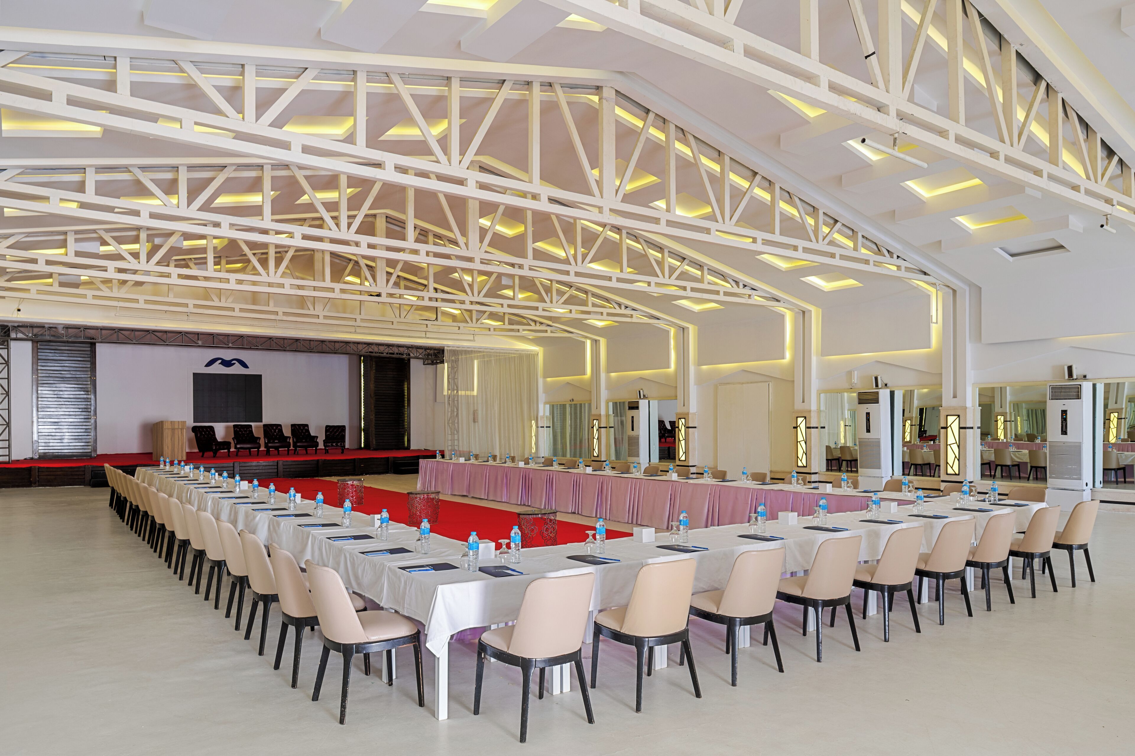 A large conference room with long tables and chairs, a stage, and a high ceiling with structural beams.