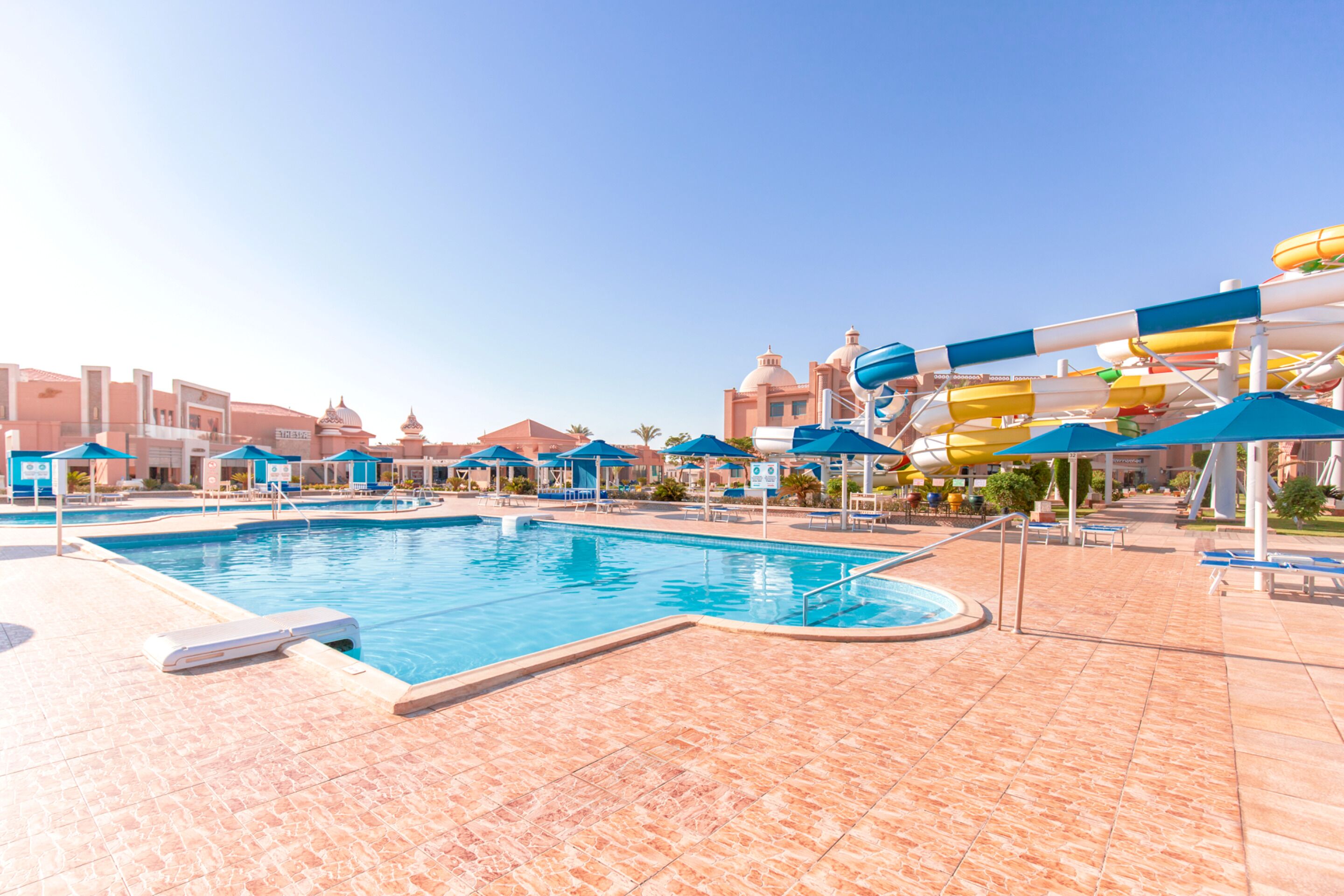 A swimming pool with water slides and umbrellas, surrounded by lounge chairs and a tiled deck.