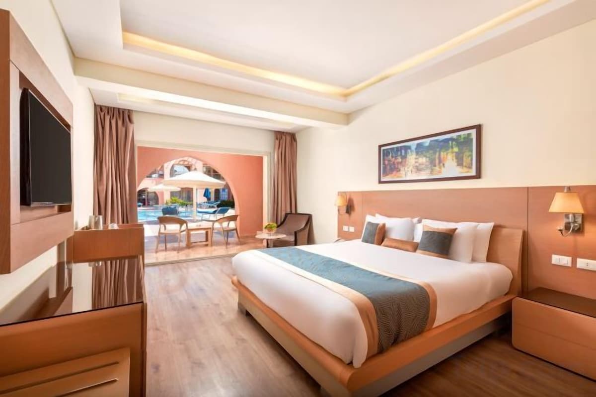 A hotel room with a large bed, a desk, a chair, a TV, and a view of an outdoor area.