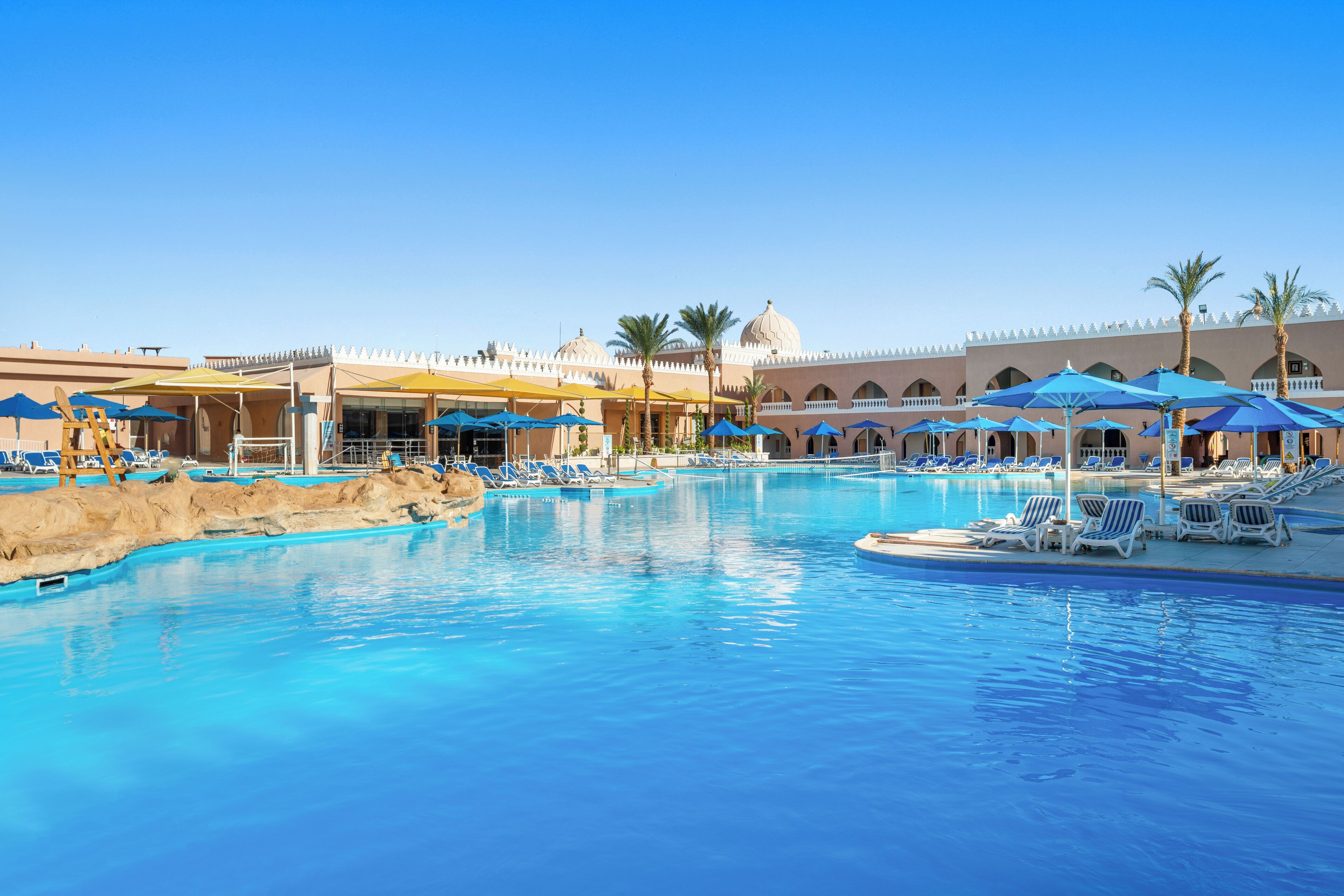 A large swimming pool with blue water, surrounded by lounge chairs and umbrellas, and a building with arches and domes in the background.