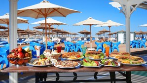 A table set with various dishes and a bowl of fruit, surrounded by blue chairs and umbrellas at a beachside location.