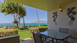 A patio with a glass table and chairs, a view of the ocean, and decorative wall art. - Royal Mauian - Maui Condo & Home (Kihei)
