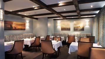 Um restaurante com mesas e cadeiras, vigas de madeira no teto e luminárias fixadas na parede.
