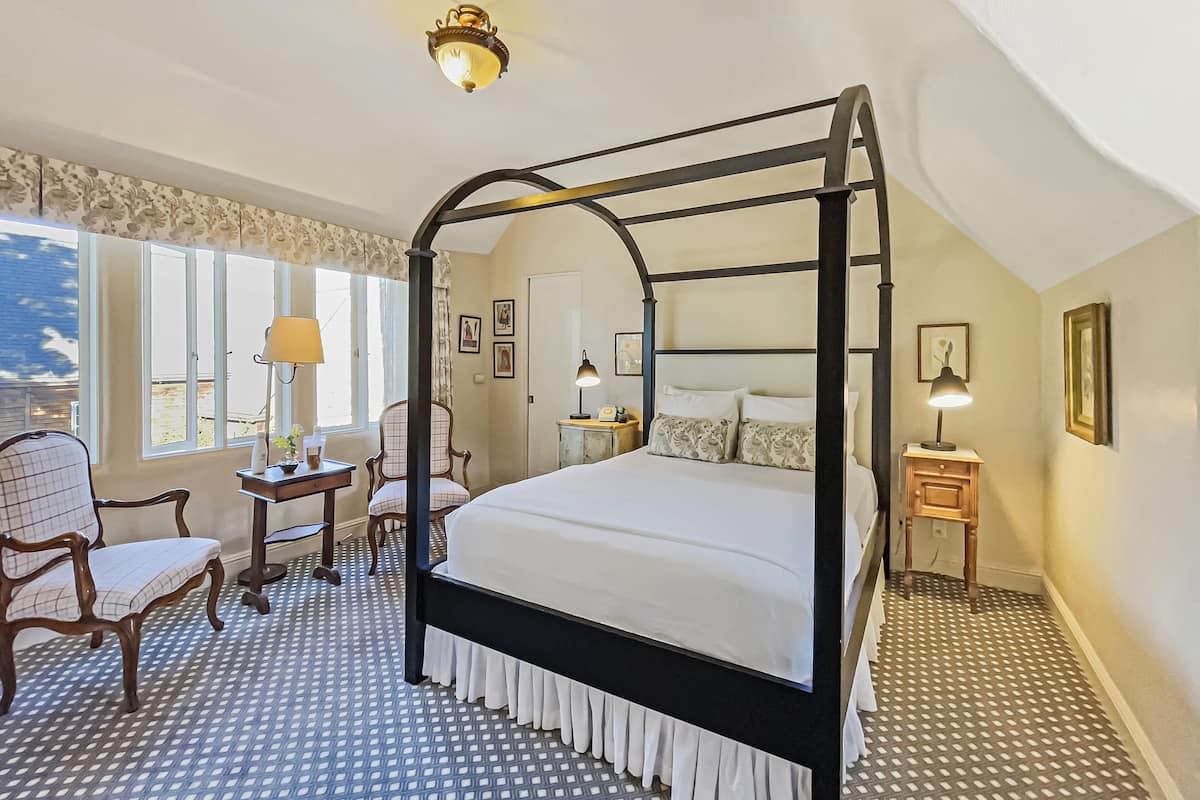 A four-poster bed with a black frame and white bedding, a desk with a lamp, a chair, and a small table with a lamp.