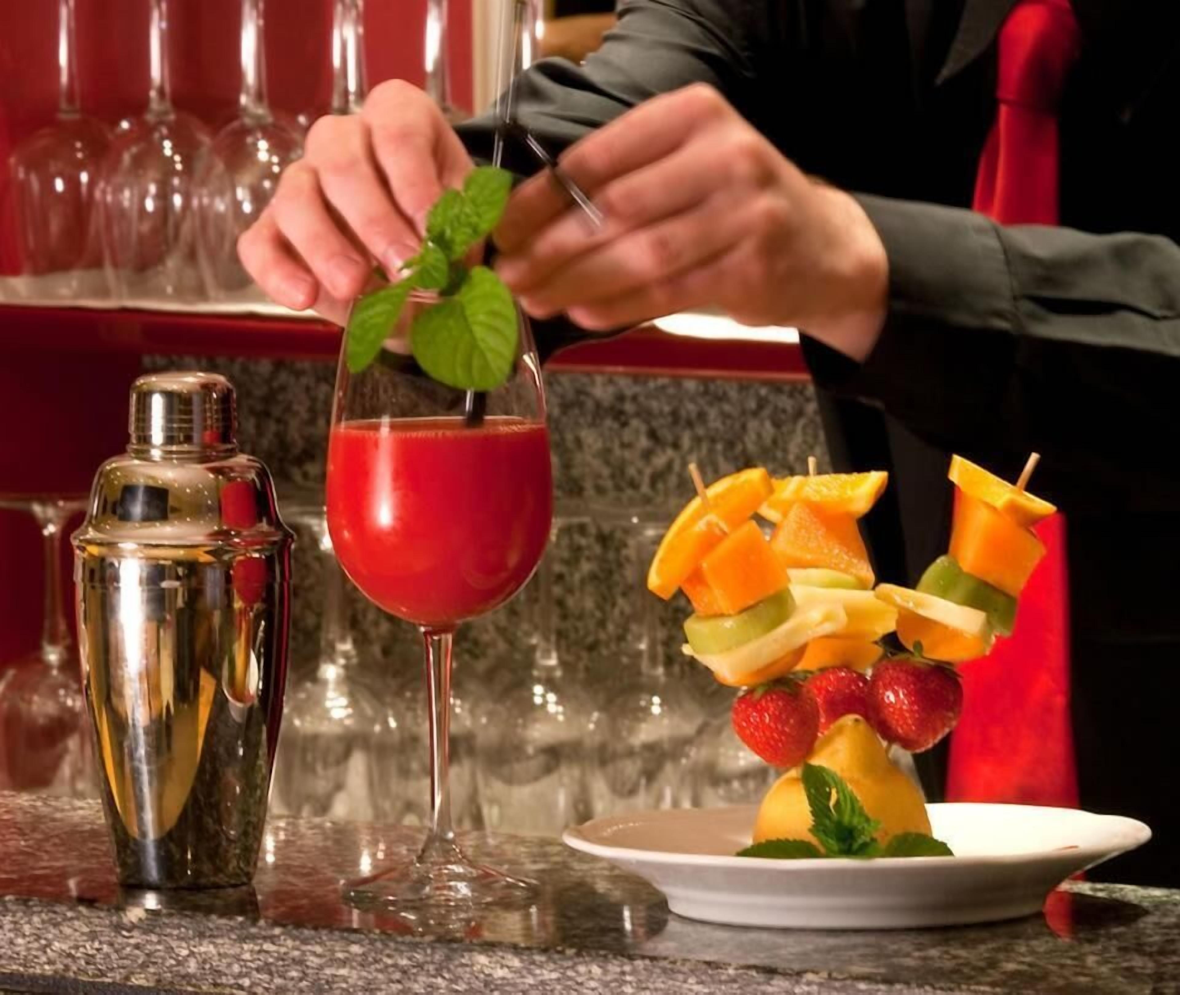 A bartender preparing a red cocktail with a mint garnish, a fruit skewer, and a cocktail shaker on a bar counter.