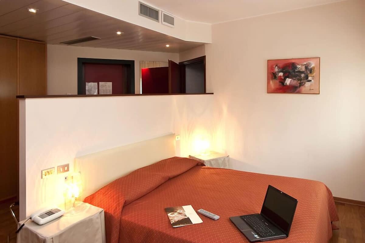 Ein Schlafzimmer mit einem Bett, einem Laptop, einem Nachttisch, einem roten Teppich und einem Bild an der Wand.