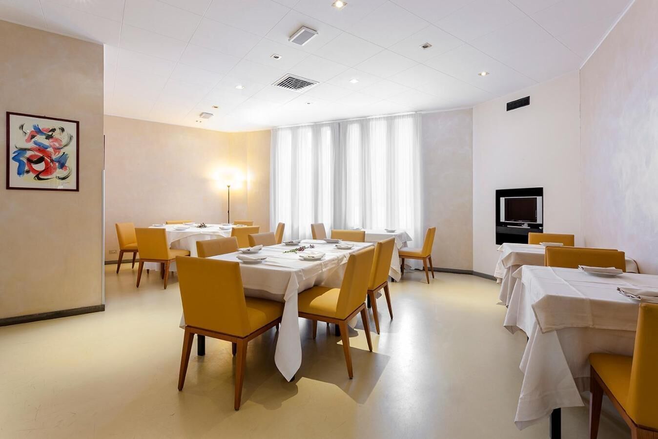 Une salle de conférence avec des tables et des chaises, une grande fenêtre avec des rideaux, un tableau accroché au mur et un téléviseur fixé au mur.