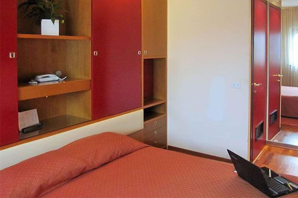 Ein Hotelzimmer mit Bett, einem Laptop auf dem Bett, einer roten Wand, einem Holzregal mit Pflanze und einem Spiegel, der das Zimmer reflektiert.
