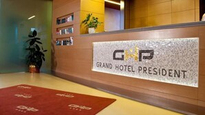 A reception area with a sign reading "Grand Hotel President" and a logo. There are bookshelves, a potted plant, and a red carpet with the hotel logo. - Grand Hotel President (Spilimbergo)