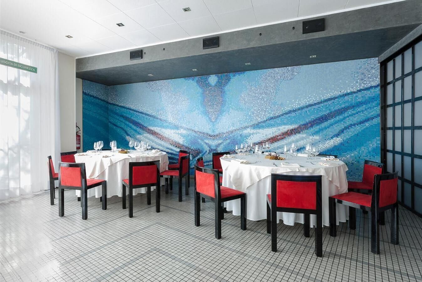 A dining area with round tables, red chairs, and a blue mosaic wall.