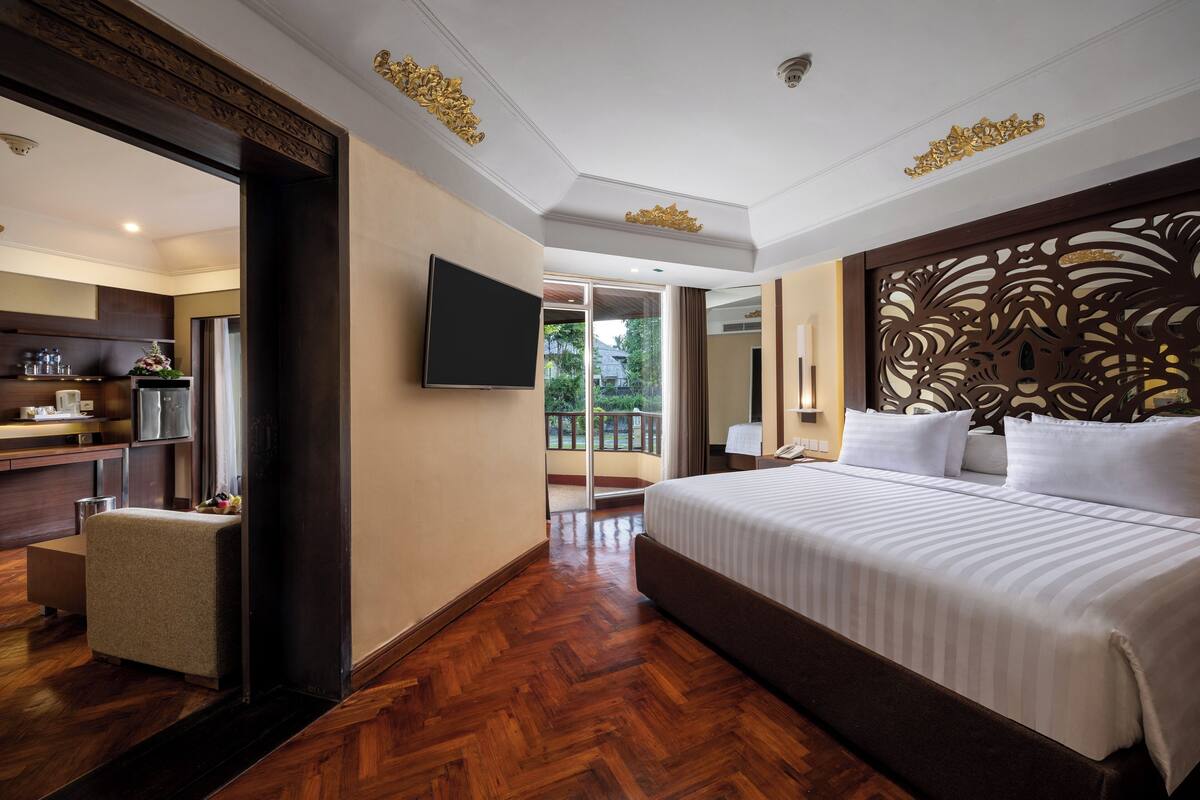 A hotel room with a large bed, a flat-screen TV, a wooden floor, and a balcony with a view of greenery.