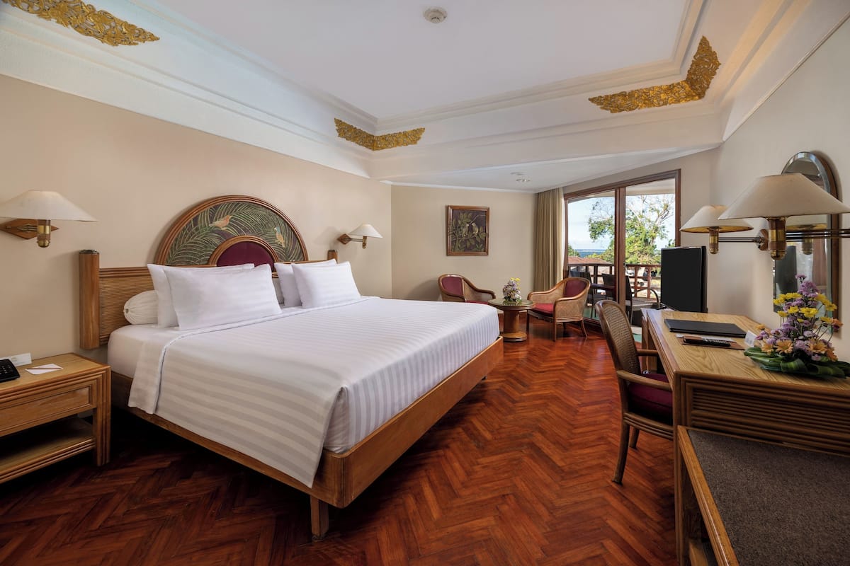 A hotel room with a large bed, wooden headboard, two bedside tables with lamps, a desk with a chair, a small table with flowers, and a large window with a view.