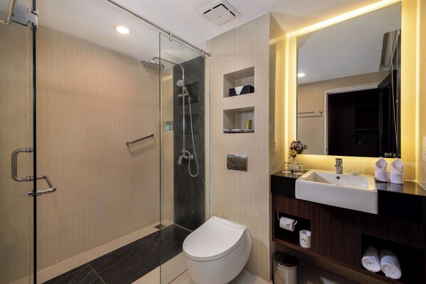 A modern bathroom with a glass-enclosed shower, a white toilet, a sink with a mirror, and shelves with toiletries. - Prama Sanur Beach Bali (Denpasar)