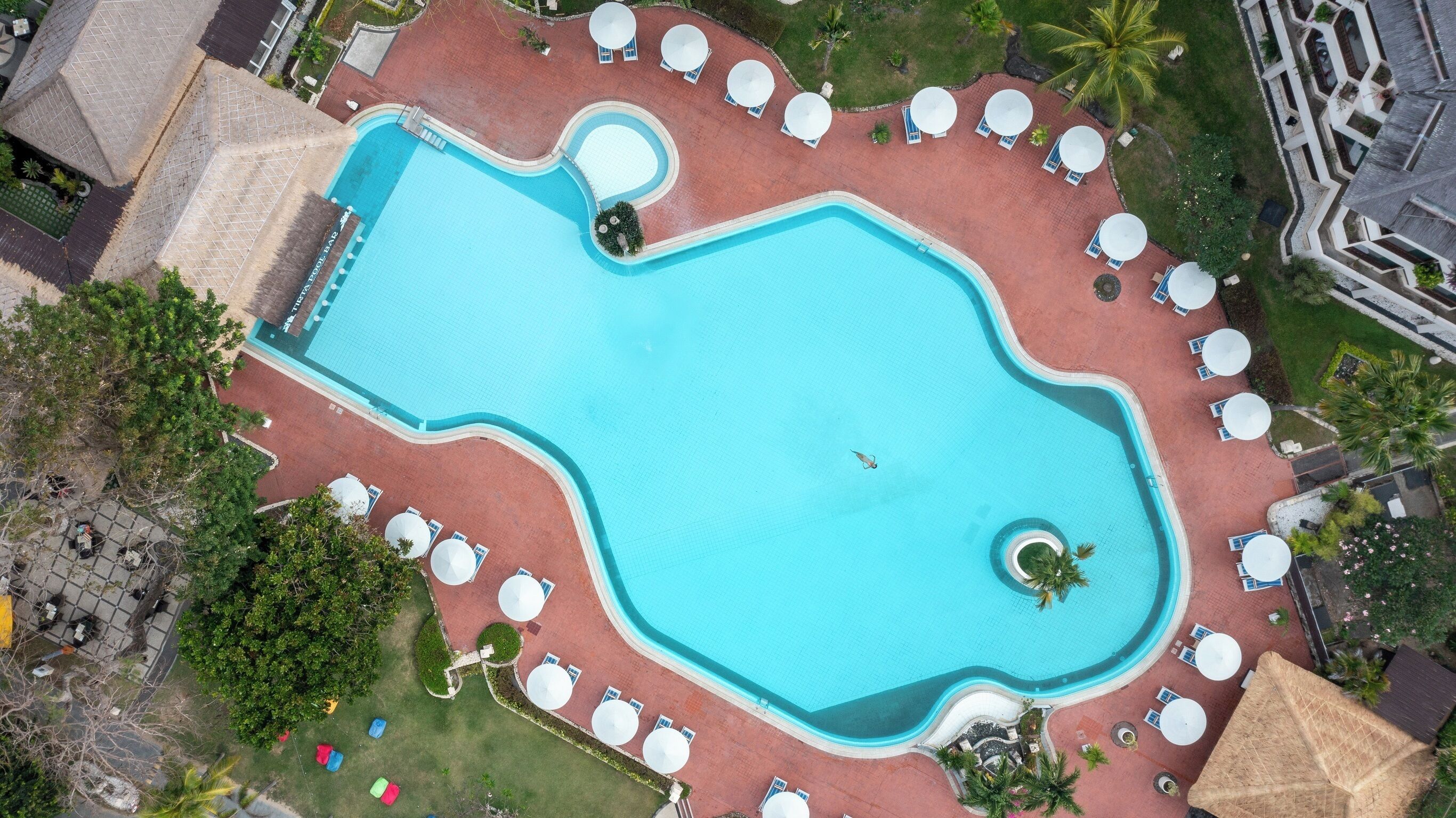 An aerial view of a swimming pool with a unique shape, surrounded by lounge chairs and umbrellas, and a small circular hot tub area.