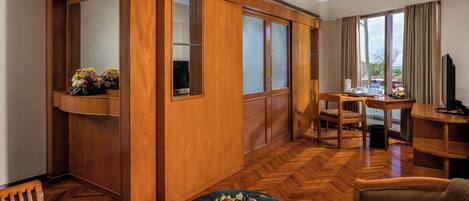 A hotel room with a wooden wardrobe, a dining area with a table and chairs, a round coffee table with fruit, and a large window with curtains.