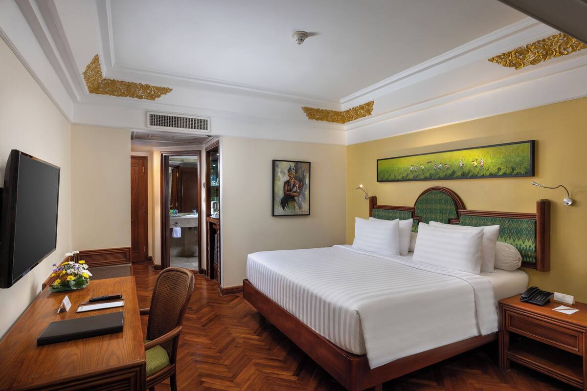 A hotel room with a large bed, a desk with a computer, a chair, a television, and a painting on the wall.