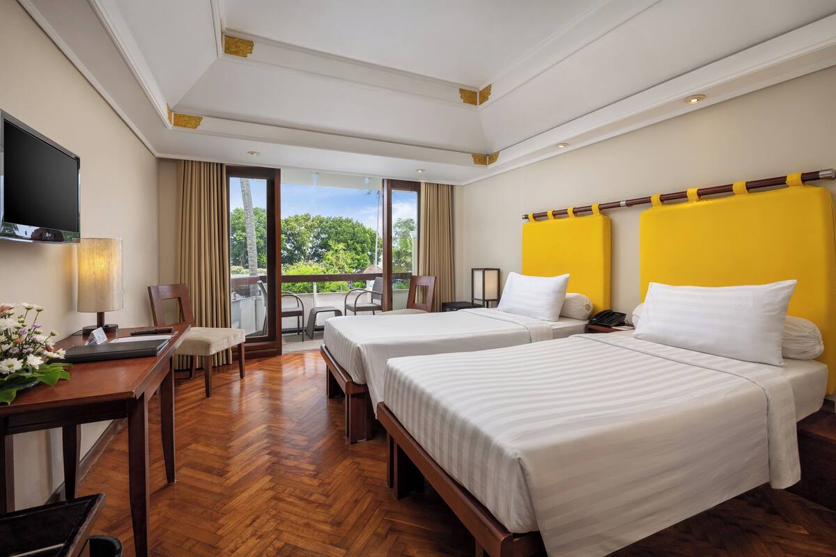 A hotel room with two beds, a desk with a lamp, a TV, and a balcony with a view of trees.