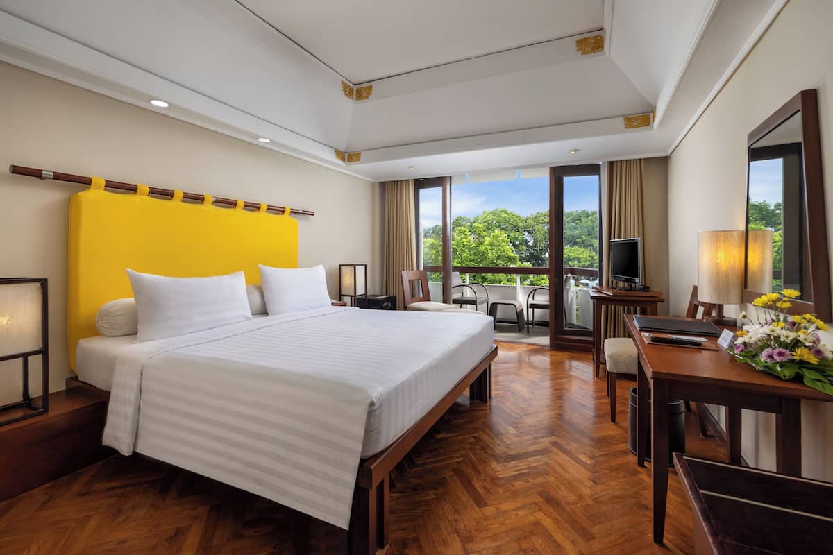 A hotel room with a large bed, a desk with a chair, a television, and a balcony with a view of trees.