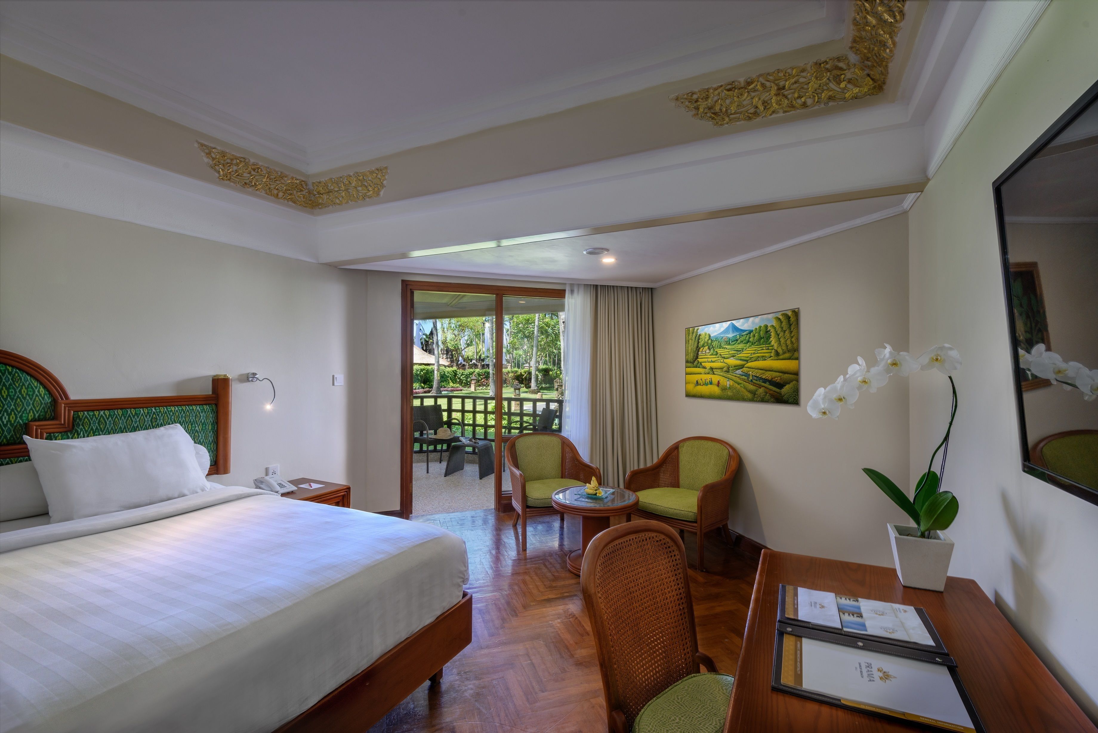 Habitación de hotel con cama, dos sillas, una mesita, un escritorio con papeles, una planta en maceta y un cuadro en la pared.
