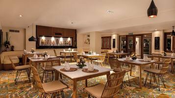 A hotel dining area with multiple tables and chairs, a large wall-mounted TV, and a patterned floor.
