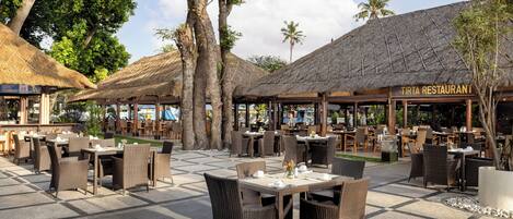 A restaurant with outdoor seating, thatched roofs, and tropical trees.
