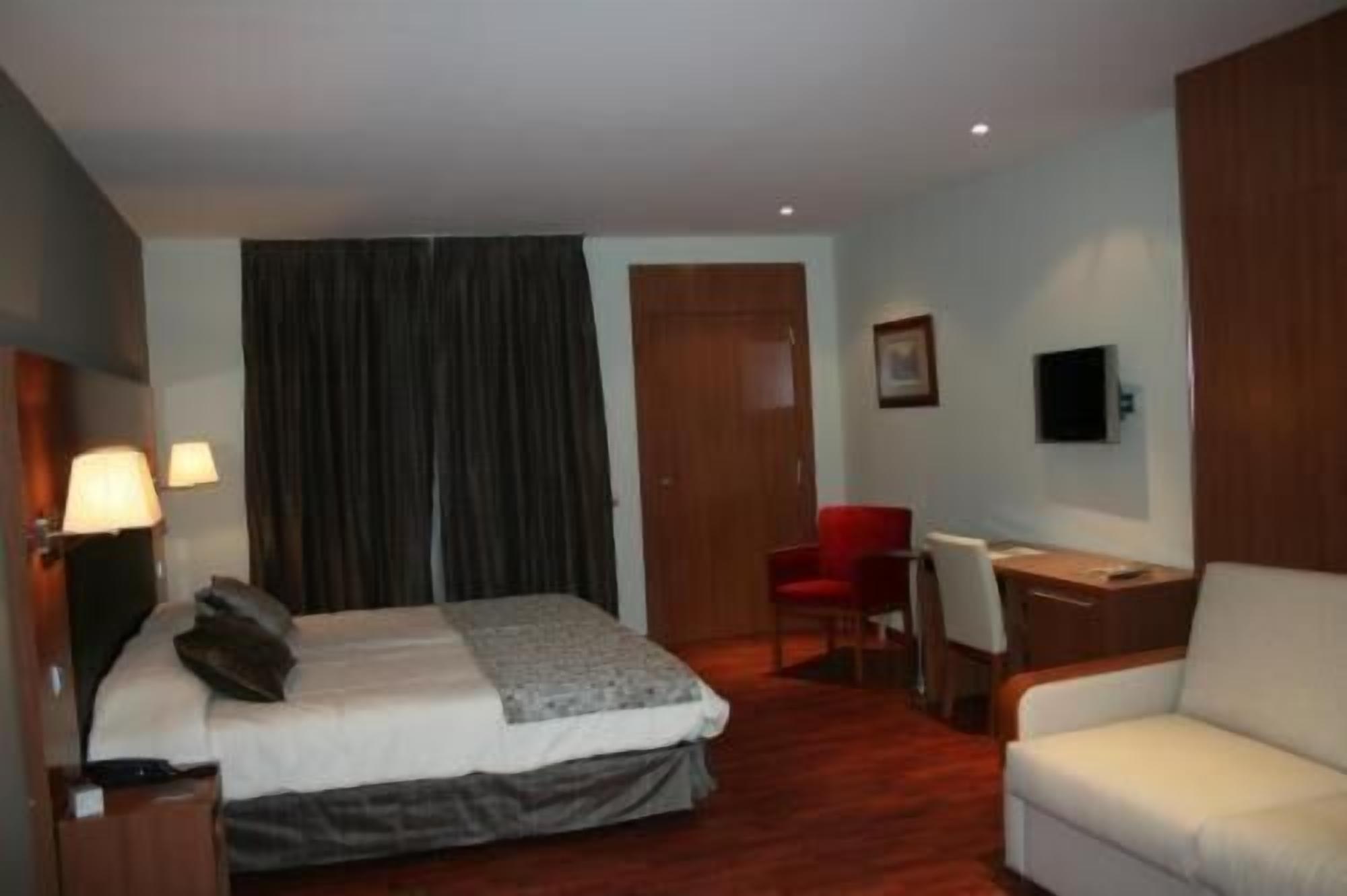Quarto de hotel com cama, escrivaninha, cadeira, televisão e janela com cortinas.