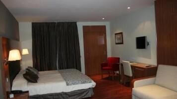Quarto de hotel com cama, escrivaninha, cadeira, televisão e janela com cortinas.
