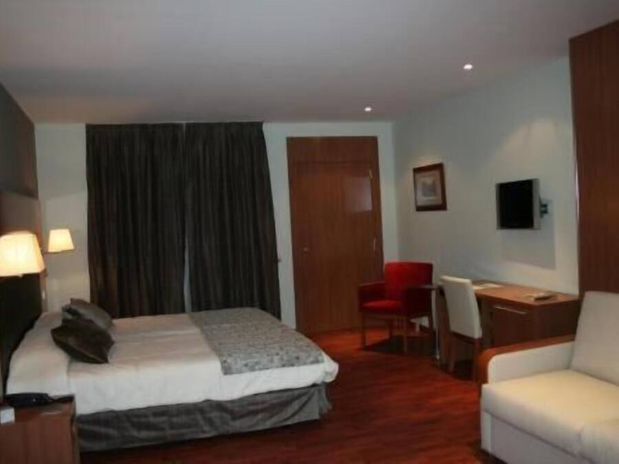 Quarto de hotel com cama, escrivaninha, cadeira, televisão e janela com cortinas.