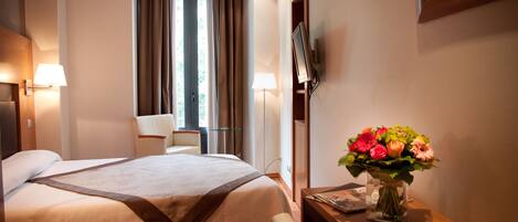 A hotel room with a bed, a desk with a vase of flowers, a chair, and a TV mounted on the wall.